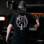 A blacsmith wearing a black t shirt with a grey and white blacksmith viking warrior holding a hammer, standing behind a anvil with red flames around border