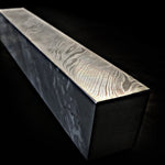 A rectangular block of Damascus steel with a highly polished top surface showing intricate twist pattern, set against a solid black background.