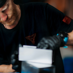 blacksmith wearing black t shirt with a red lava vegas forge logo on the chest and red american flag on left sleeve. he is stacking layers of steel for damascus steel canister