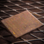 a ladder pattern mokume flat bar with layers of copper, brass and nickel-silver sitting on a metal corrugated background