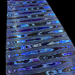 A close-up of a patterned zirmascus flat bar with horizontal wavy bands in electric blue, violet, black, and metallic silver, shown against a black background.