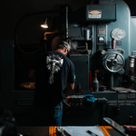 a man wearing a black vegas forge war hammer graphic t shirt while he is running a blanchard surface grinder
