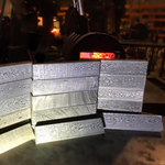 A group of rectangular damascus steel billets or ingots with a textured, patterned surface, arranged on a workbench in a workshop setting with a forge visible in the background.