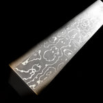 A close-up of a long, slender silver damascus steel block etched with wavy pattern, photographed diagonally against a solid black background.