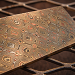 close up image of a riptide patterned mokume gane billet with layers of copper, brass and nickel-silver that flow throughout it, sitting on a metal corrugated background