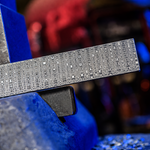 snakeskin pattern damascus billet sitting on nazel hammer sow block with blue and red toned forge in background