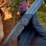 a folding knife with snakeskin pattern damascus blade being held by black glove hand with flowers in background