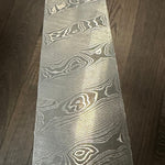 A close-up image of a twist pattern damascus steel billet with brown wood background