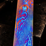A close-up of a damtanium twist flat bar with a vibrant, multicolored wavy pattern of blue, purple, orange, and red, set against a dark, textured forge background.